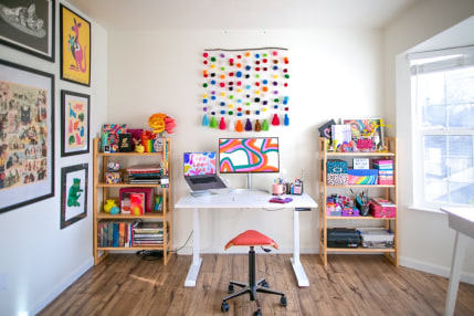 Christine's workspace features pops of color throughout. At the center is her computer setup with a kooky rainbow pom-pom wall piece hanging on the all above. Her workforce is flanked by two brightly decorated shelves. To the left is a wall of art and a wall of windows is on the right. It's a very bright, naturally lit space with light off-white walls and a warm dark wood floor.