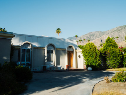 A ranch-style home against a mountain backdrop in early morning light with palm trees in the background