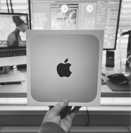 Black and white photo of me holding my new Apple Mac Mini out in front of me. In the foreground is the Mac Mini in my left hand, while the background is out of focus showing a preview of a project I'm working on.