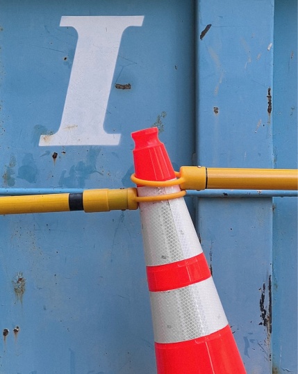 An italicized serif “I” spray painted in white on a rusty light blue metal panel with yellow poles wrapped around a blood orange traffic cone at a construction site. 🤝