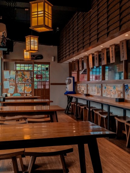 A cozy small Japanese cuisine restaurant in Tangerang Selatan, Indonesia, with wooden tables, chairs, a long counter, and sliding doors. Paper lanterns on the ceiling cast a warm glow.