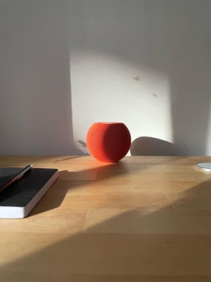 A centered orange HomePod mini sitting on a wooden desk with some natural sunlight over it.