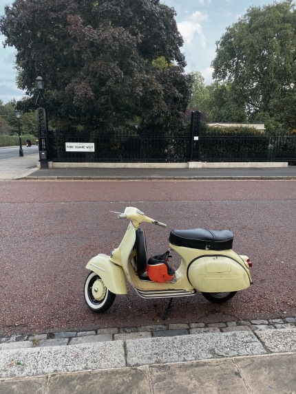 This photograph is of an old Vespa, that is parked at the end of a residential street called 'Park Square West', in North West London. Its yellow paint is beginning to discolour, and it is slightly chipped around some parts of the scooter - further showing its age. The owner has left their orange helmet tucked at its base. The photo's backdrop is a beautiful private residential park, with vibrant green and purple trees that tower over the its black gates.