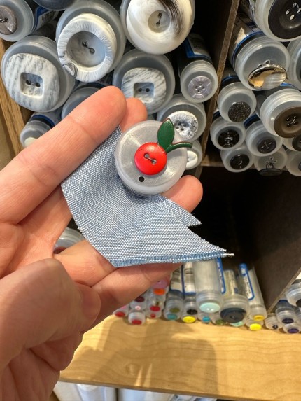 Red button with green leaf on top making it resemble an apple or cherry on top of a light
blue fabric. Other buttons in the background in tubes