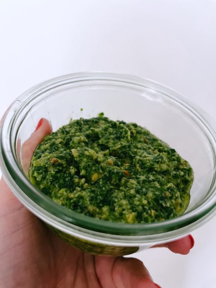 Glass jar of green pesto, made from wild garlic, held up in a left hand against a white wall.