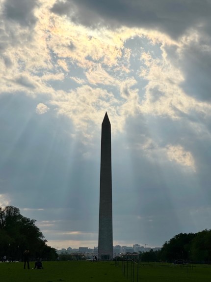 Cloudy skies parting as beams of light shine down on the Washington Monument, a white obelisk shaped building.