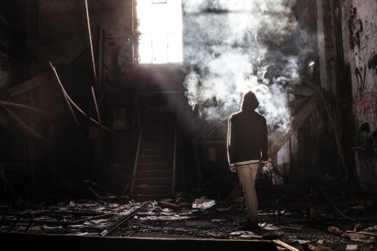 Man with a hood up standing in a destroyed house with rubble all around him.