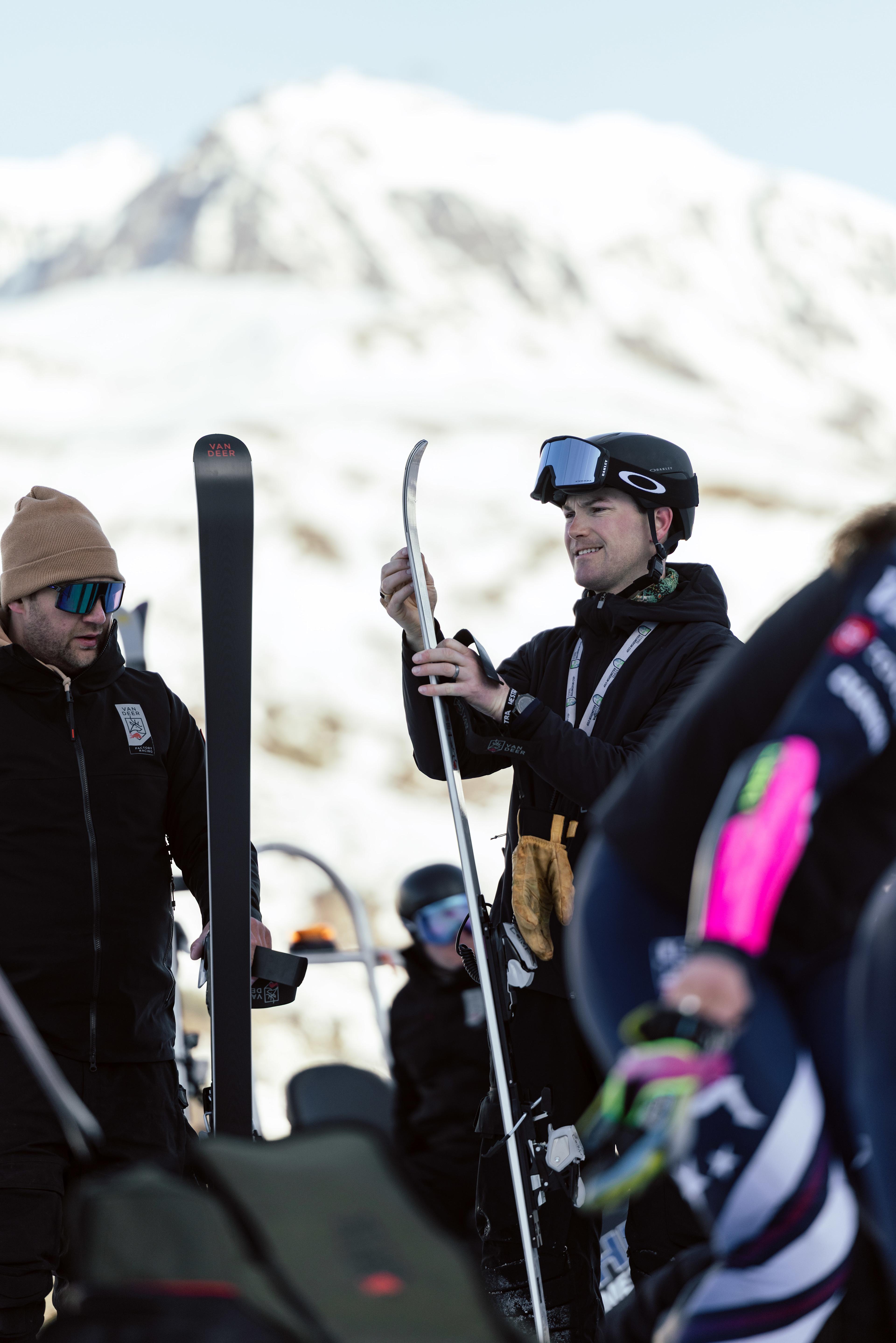 Mehrere Personen stehen auf einer verschneiten Skipiste; ein Mann mit Helm und Skibrille hält einen Ski und prüft dessen Kante.