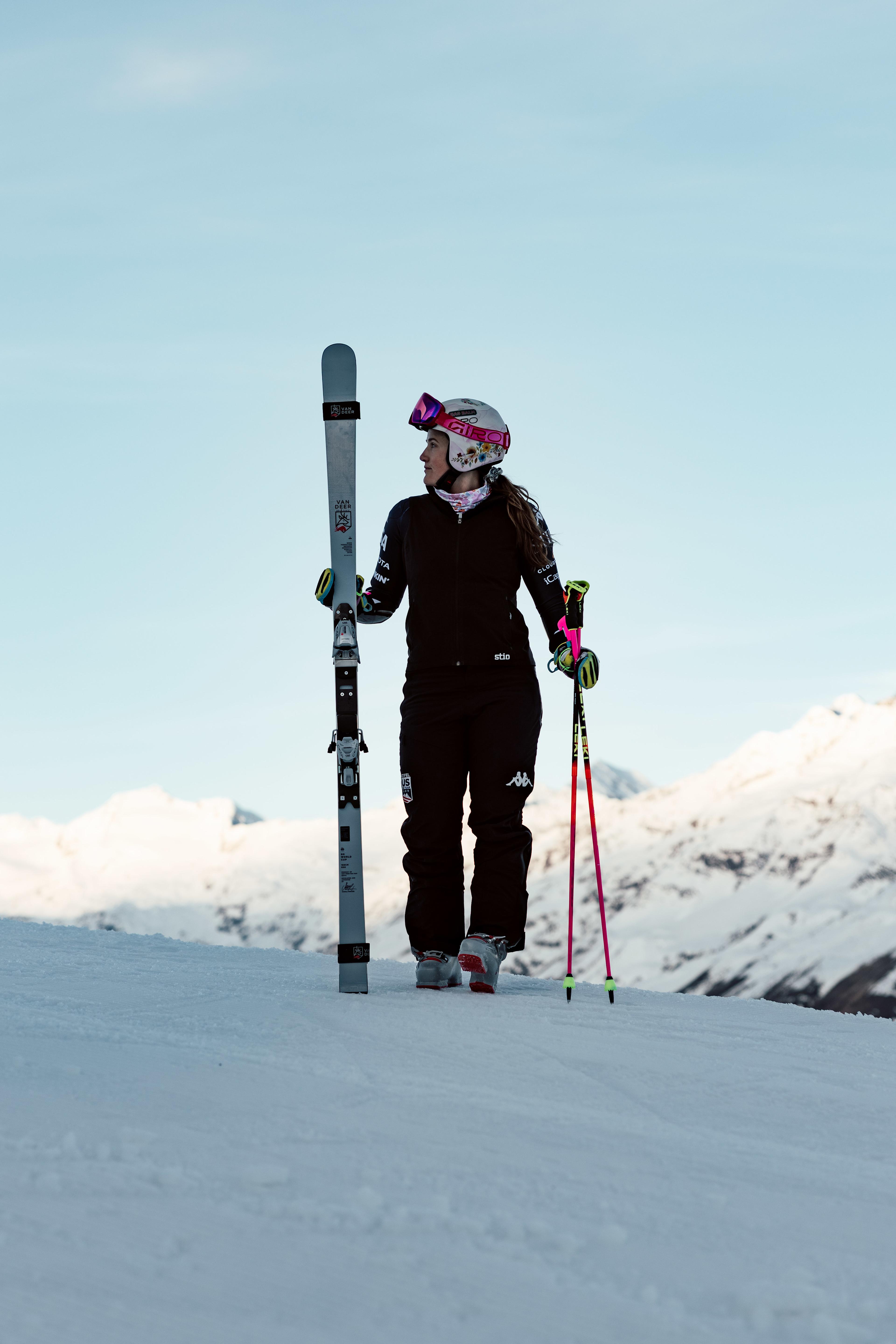 Skirennfahrerin steht auf einer verschneiten Bergkuppe, hält ihre Ski und blickt zur Seite, im Hintergrund schneebedeckte Berge.