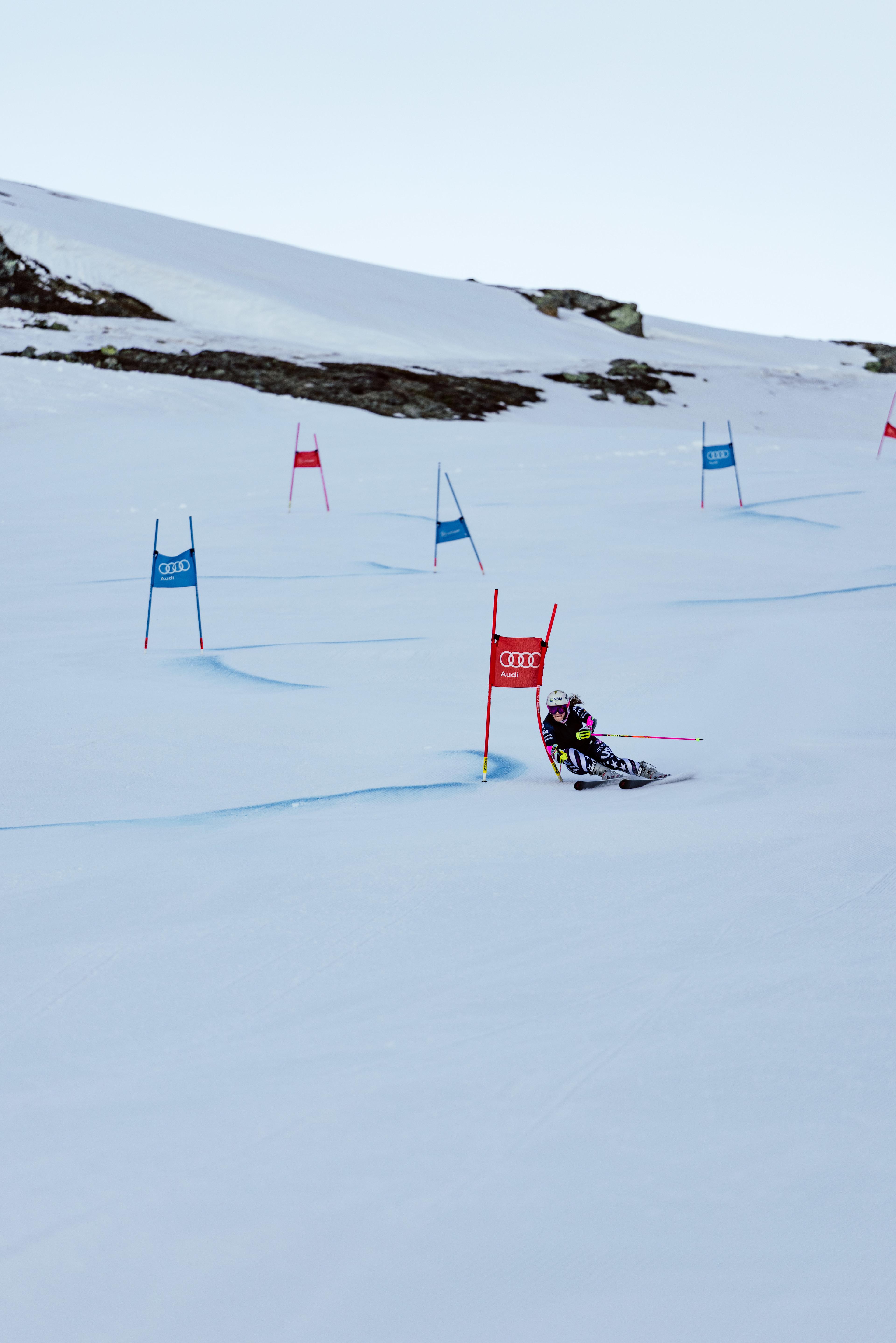 Eine Skirennläuferin fährt mit hoher Geschwindigkeit durch ein Tor auf einer verschneiten Piste. Sie lehnt sich tief in die Kurve, trägt einen Helm mit Skibrille sowie einen Rennanzug und hält Skistöcke in den Händen.