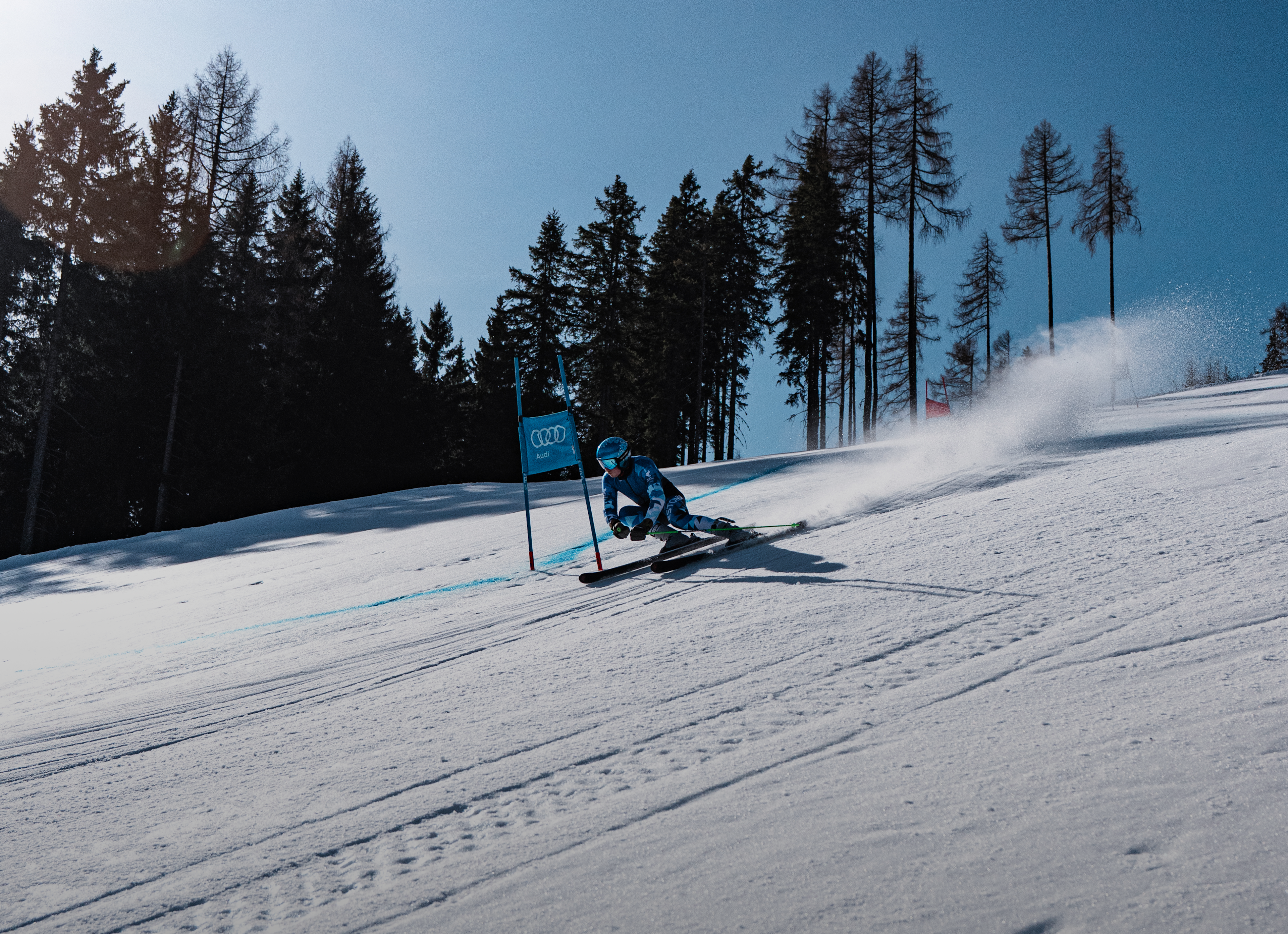 Skirennfahrer in voller Geschwindigkeit beim Riesentorlauf auf einer verschneiten Piste, er lehnt sich tief in die Kurve zu einer blauen Torstange.