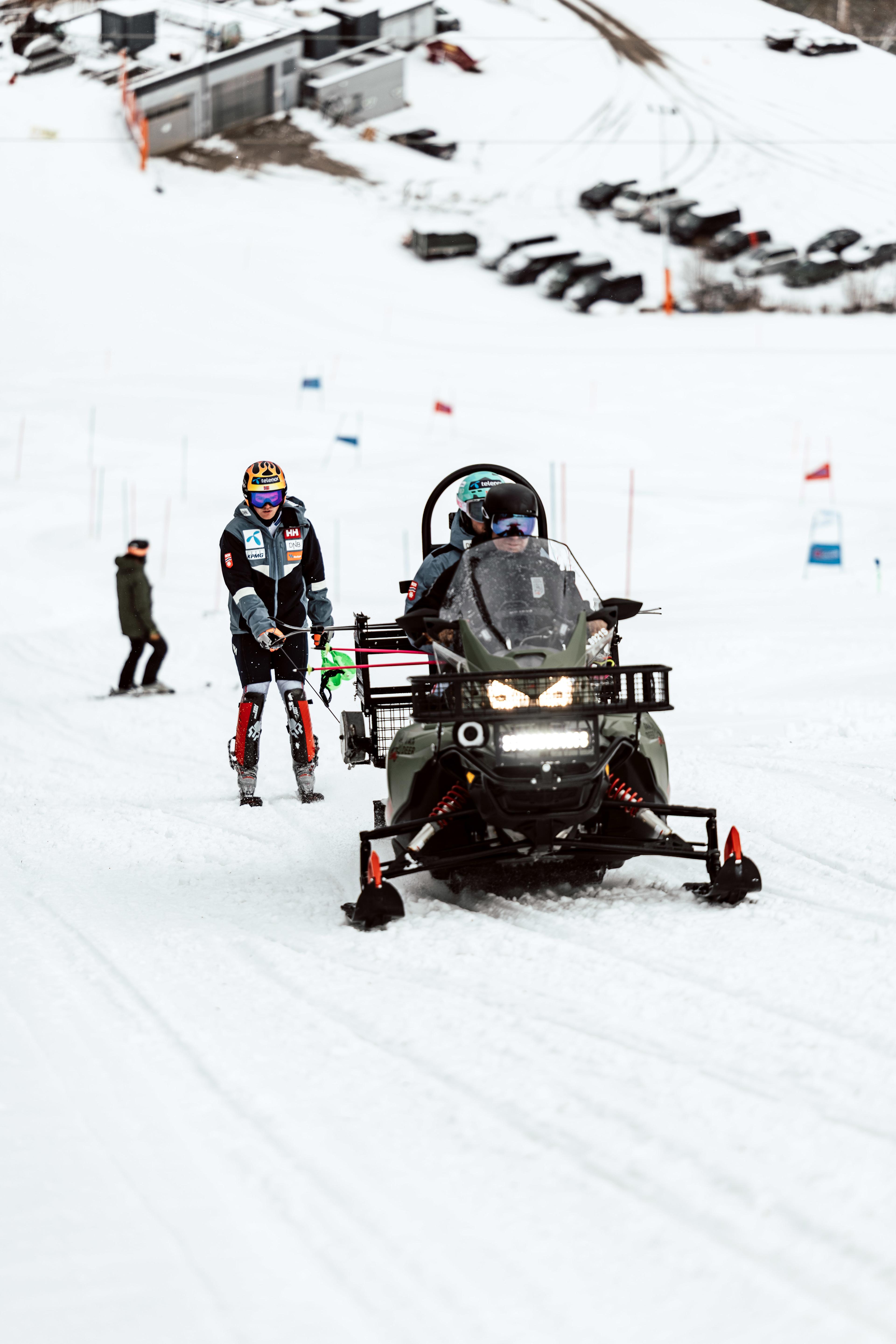 A ski racer is being pulled uphill on skis by a snowmobile next to the ski slope