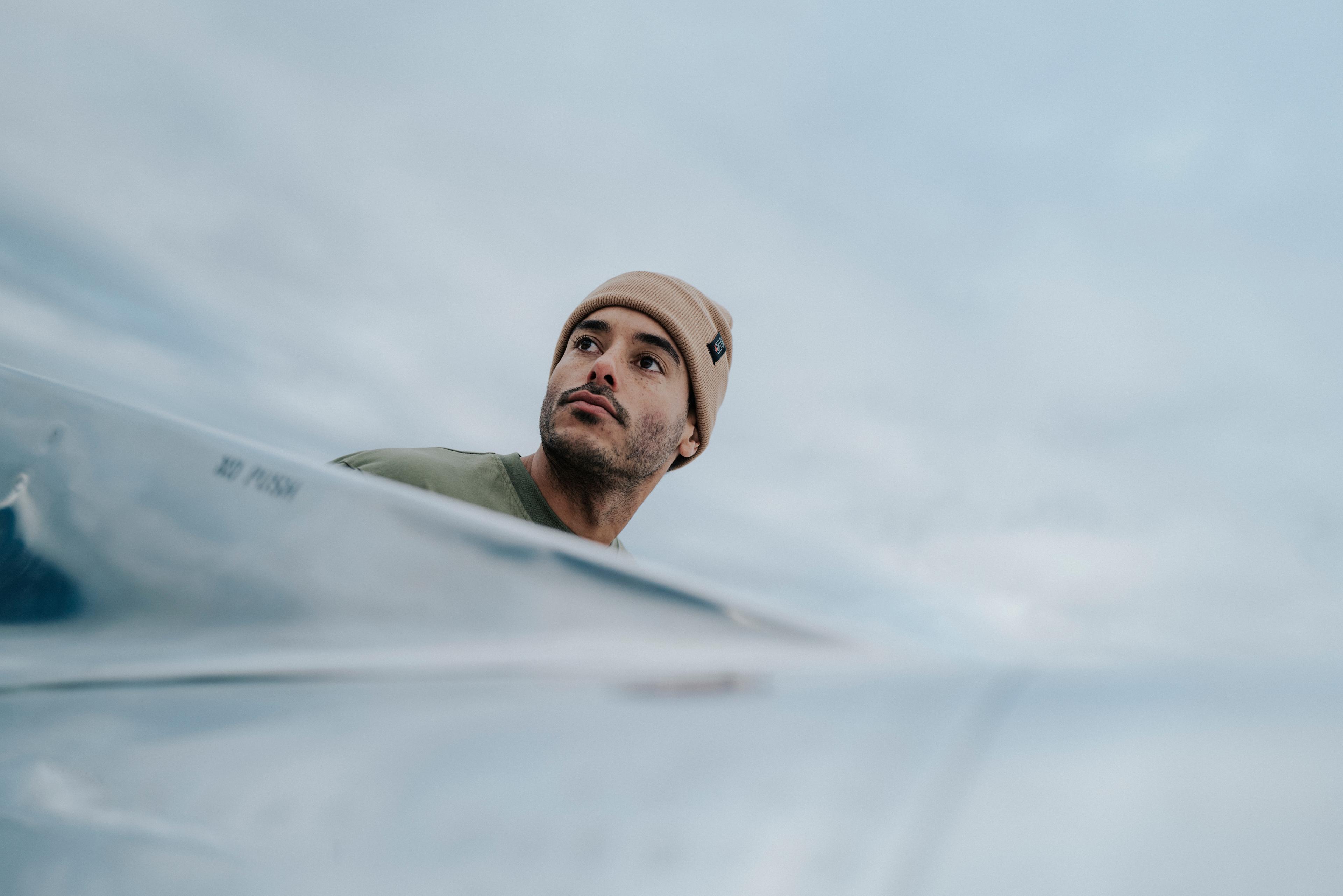 A man wearing an ocher VAN DEER-Red Bull Sports beanie is looking to the side. The close-up focuses on the beanie and the man’s side profile.