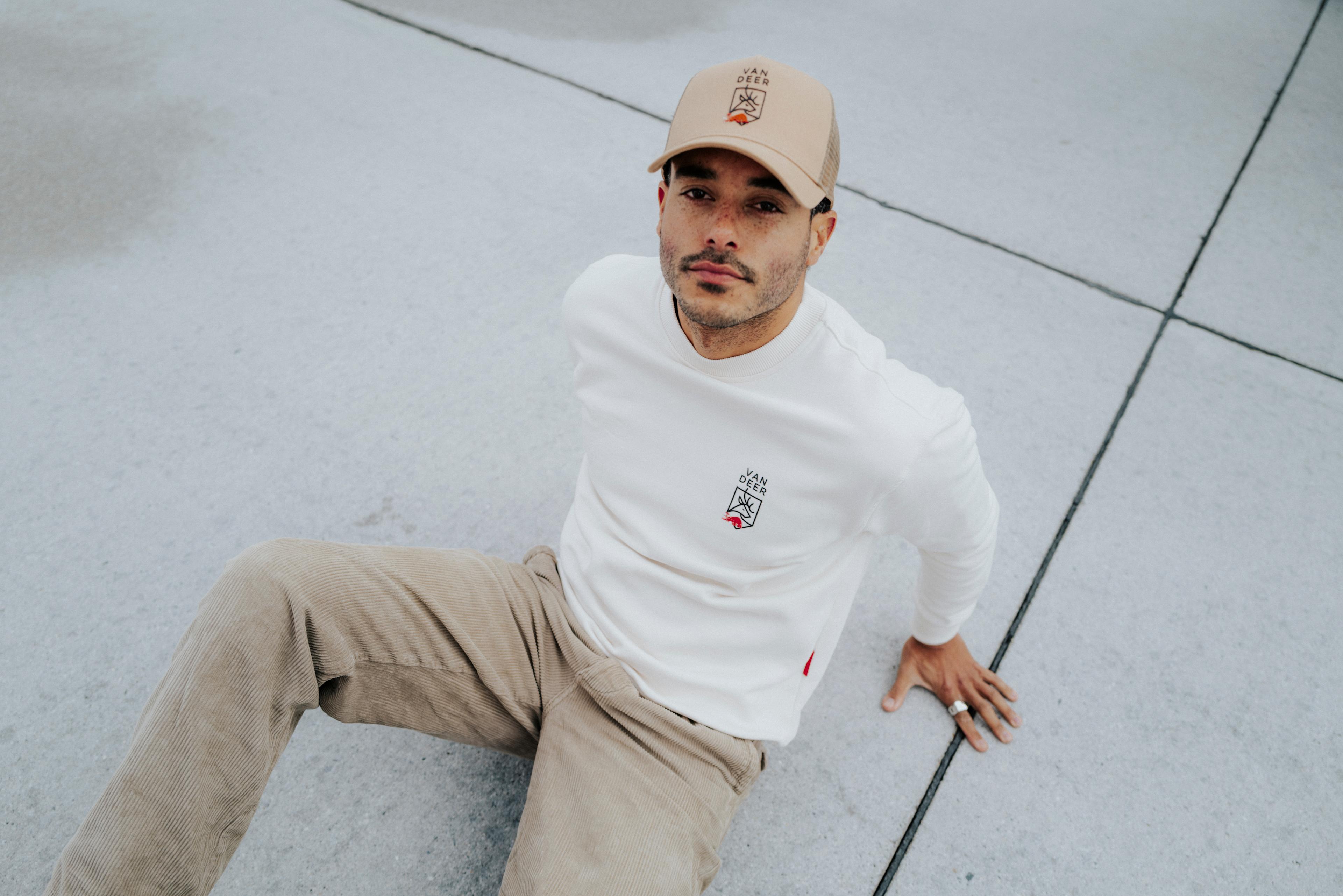 Man sitting on the ground wearing an off-white VAN DEER-Red Bull Sports long-sleeve T-shirt with a chest logo and a matching beige VAN DEER-Red Bull Sports trucker cap, paired with beige corduroy trousers.