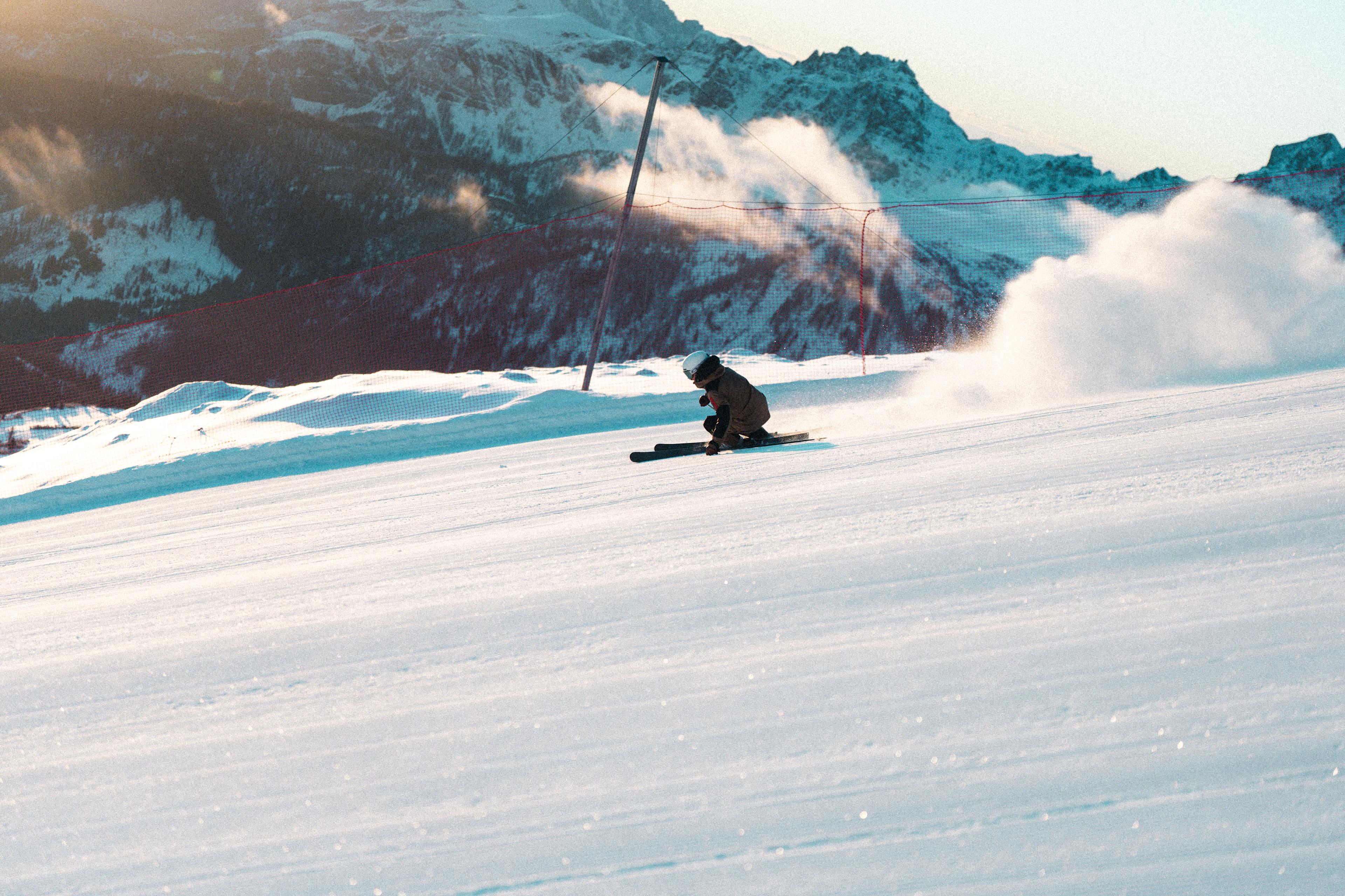 Ein Skifahrer auf VAN DEER-Red Bull Sports PRO Skiern fährt in tiefer Vorwärtshocke eine präparierte Piste in Alta Badia hinunter. Im Hintergrund sind schneebedeckte Gipfel zu sehen. Die Szene ist in warmem Gegenlicht getaucht.