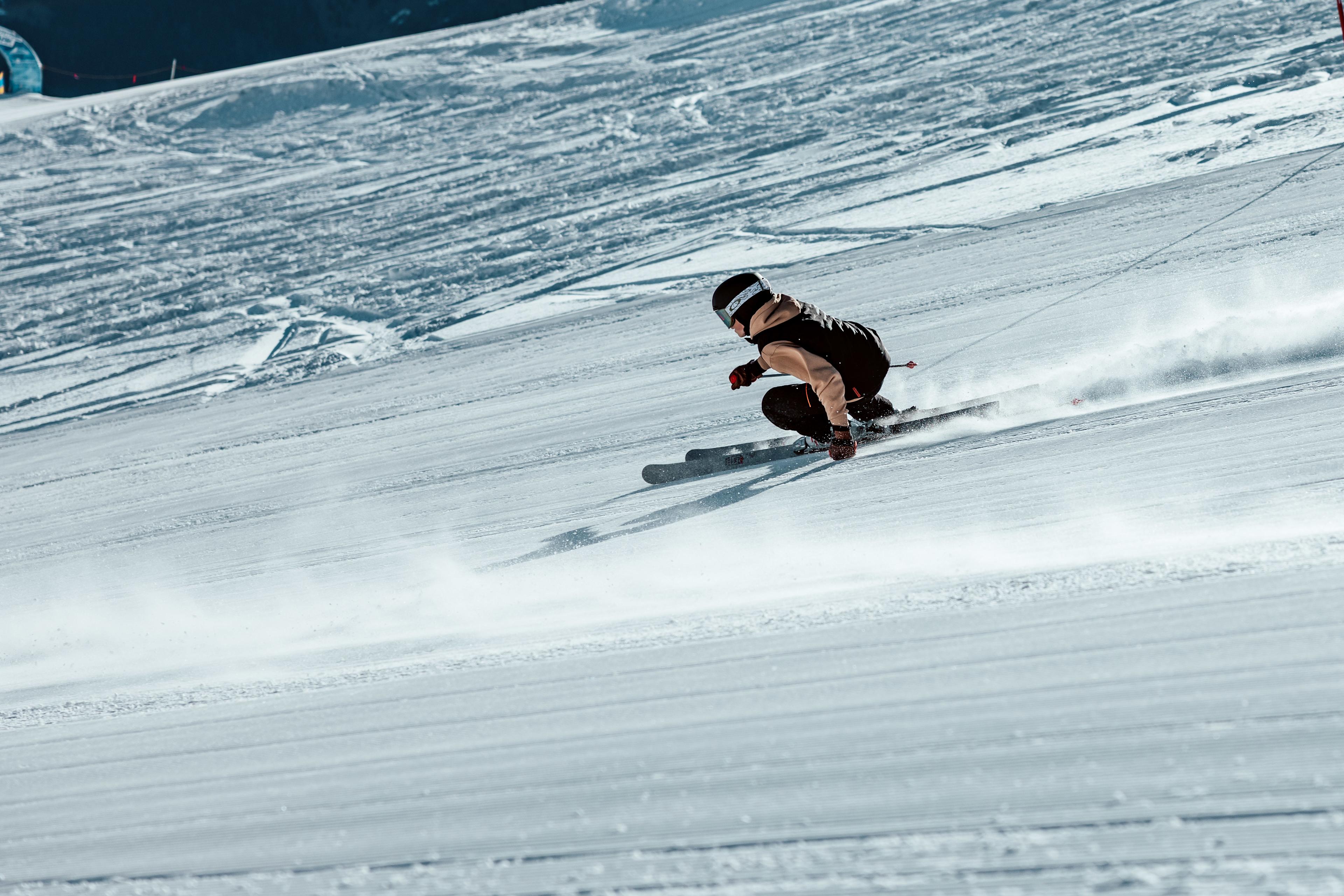 A young skier in a black race suit descends a steep, sun-lit slope at high speed on VAN DEER-Red Bull Sports Junior Pro Series skis, spraying a trail of snow behind them.