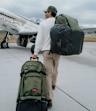 Traveler with VAN DEER–Red Bull Sports x OGIO luggage in front of a vintage airplane in an alpine setting.