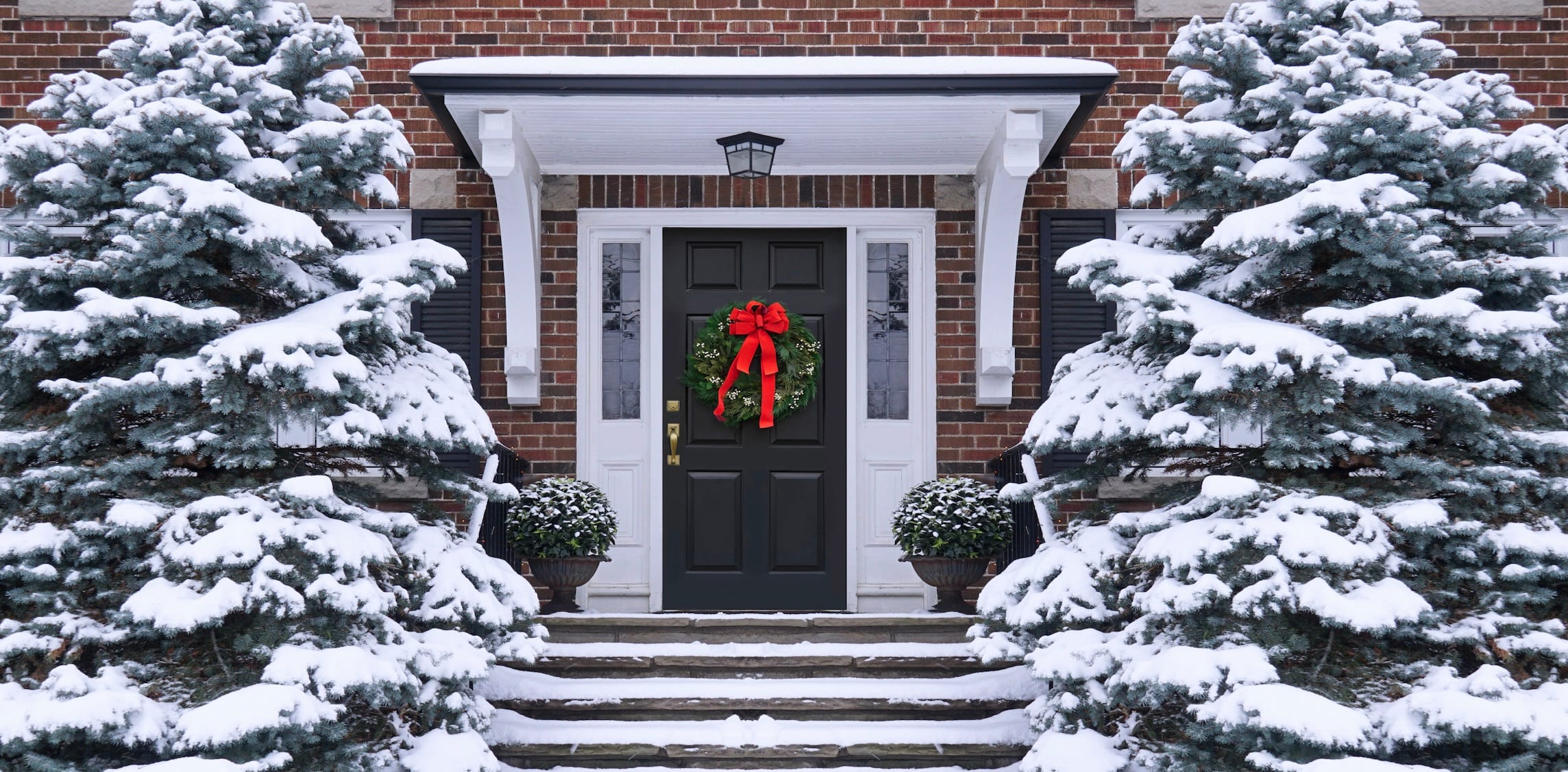 Holiday wreath hanging on the front door of a brick home with snowfall in the yard.