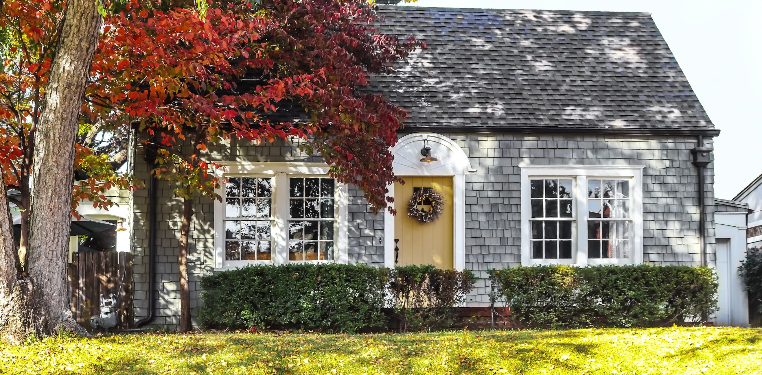 Grey house with a yellow door and trees in the grassy front yard.
