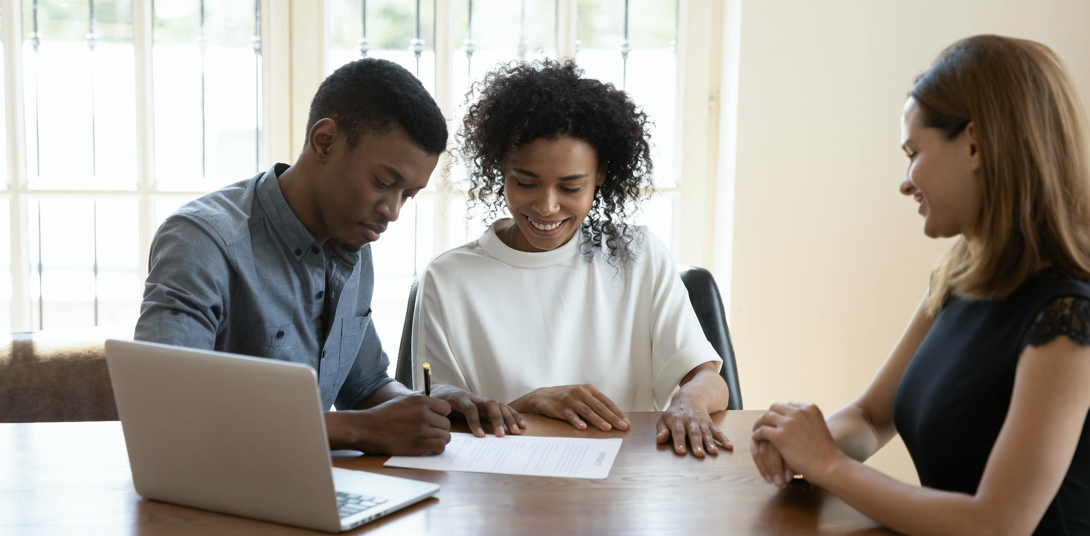 Two people meet with a professional to sign papers. They are all sitting around a wood table with a silver laptop computer on it.