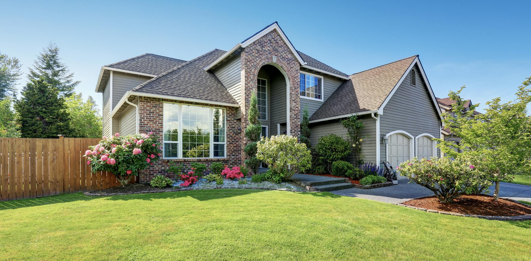 Green grass, wood fencing, and pink flowers outside of brick and grey house.