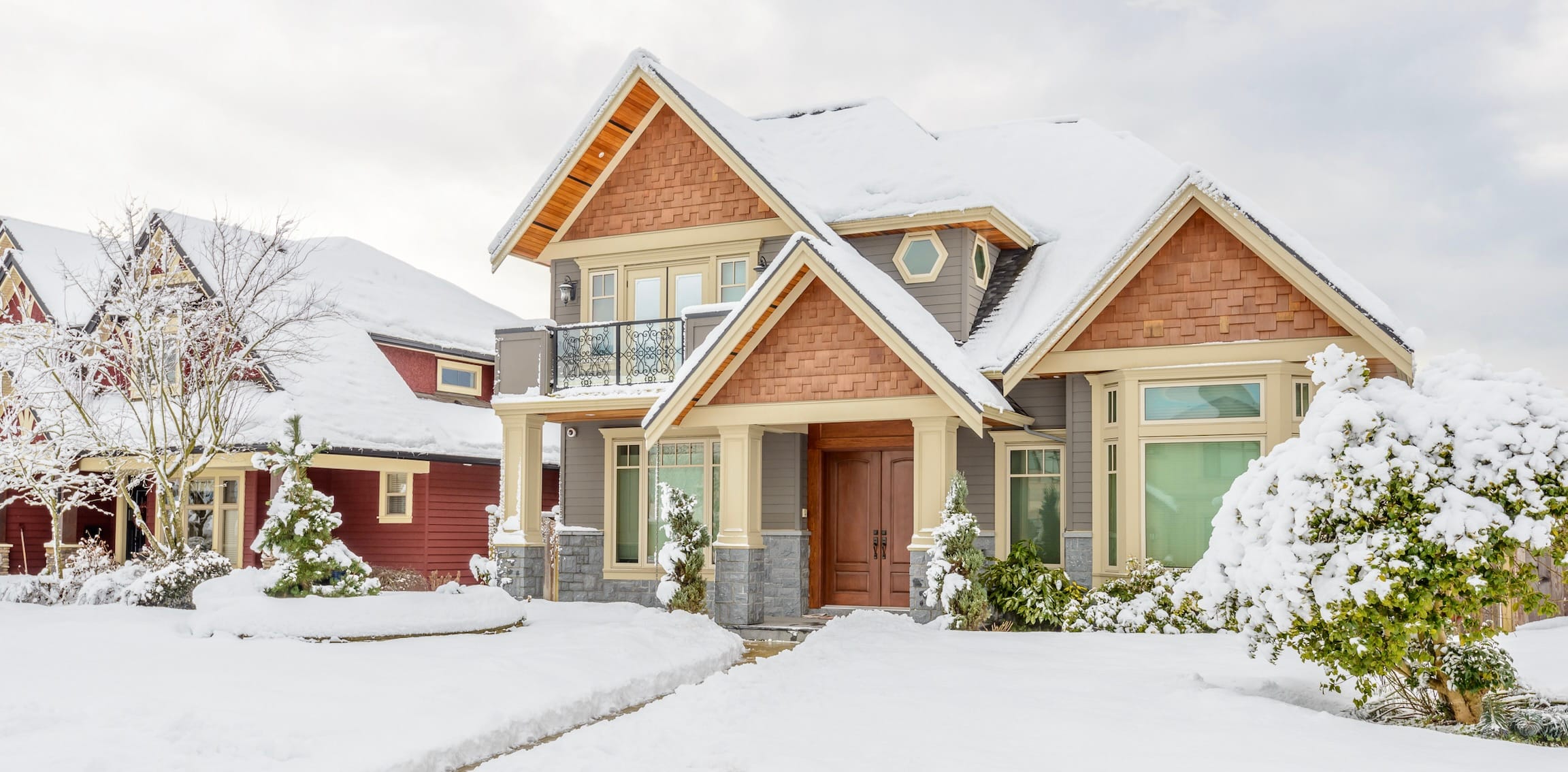 Grey and brown house with a wood front door and snow covering the yard and shrubbery.