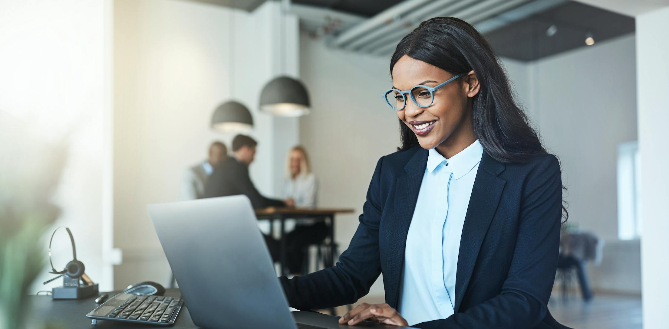 Woman wearing a blue shirt and blue glasses sitting at a desk and working on a computer.