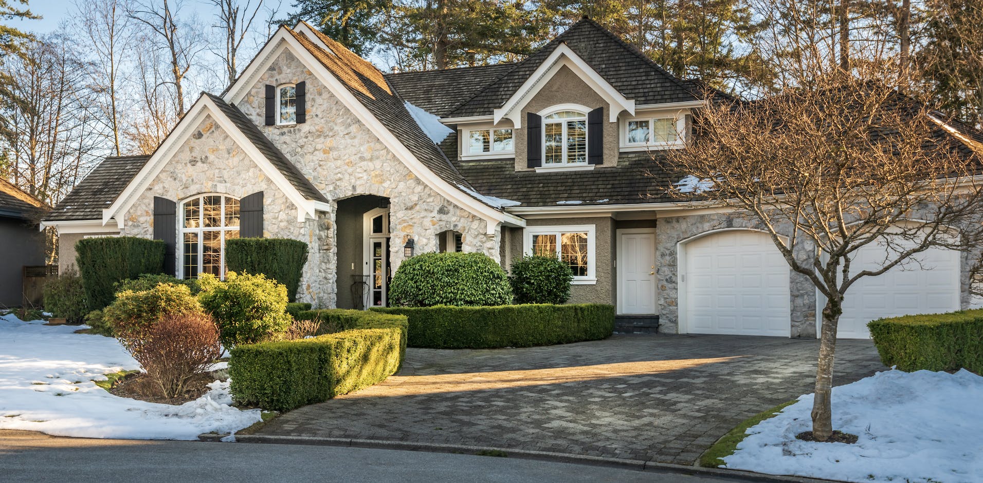 Stone home with green hedges, two white garage doors, and snow in the front yard.