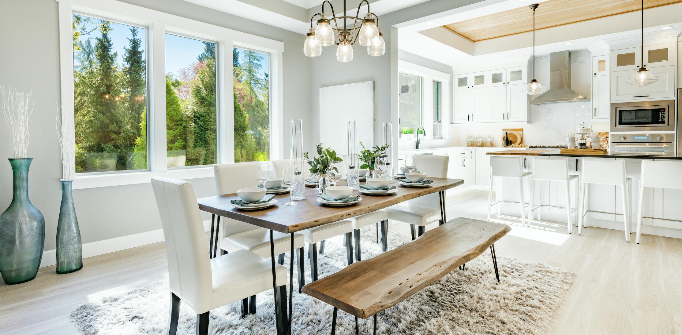 The dining room and kitchen staged with a table, chairs, rug, dining ware, vases, and a pendant light.