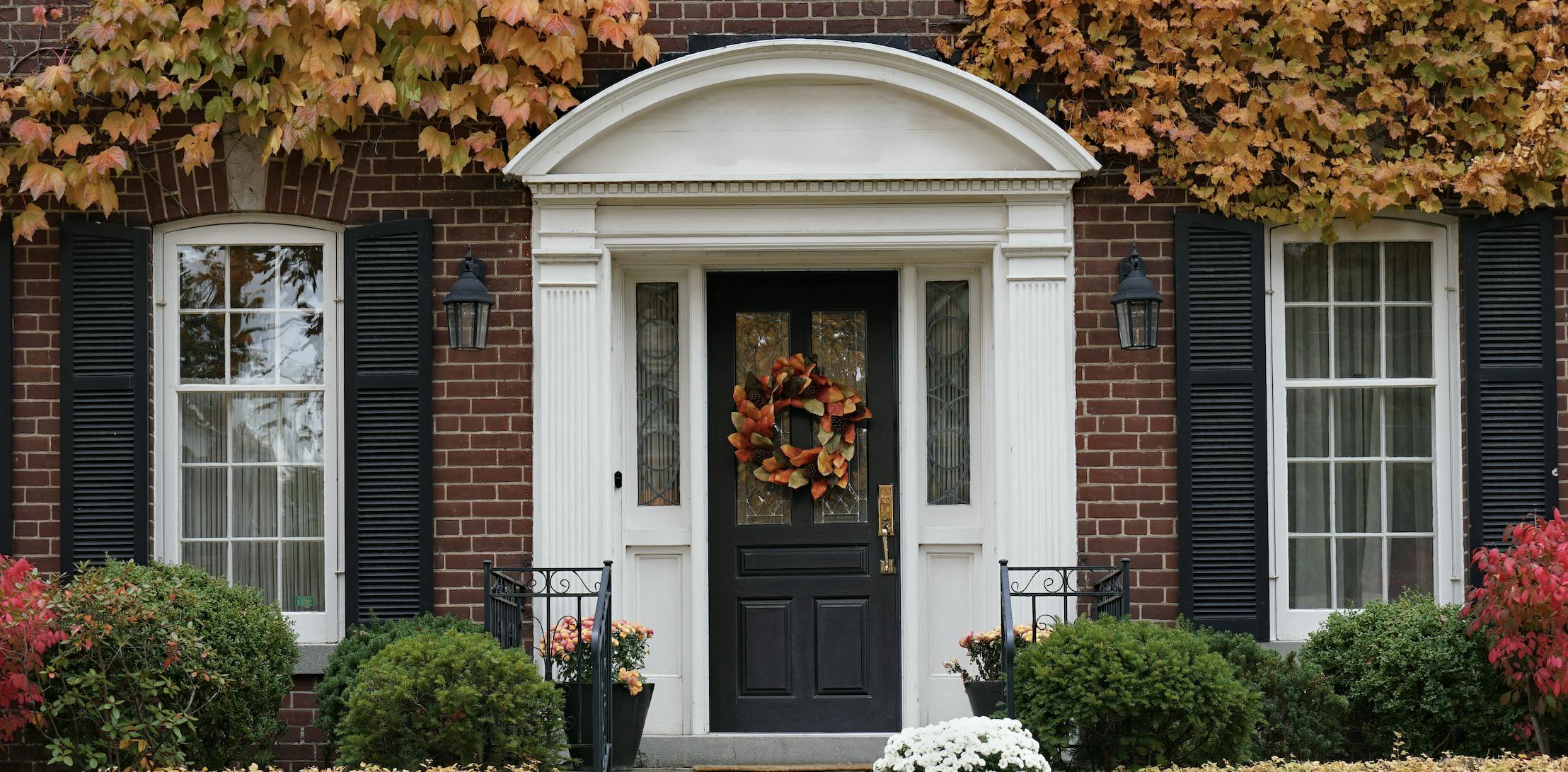 A brick home with black shutters, white time, fall foliage surrounding windows, and a seasonal wreath on the front door.
