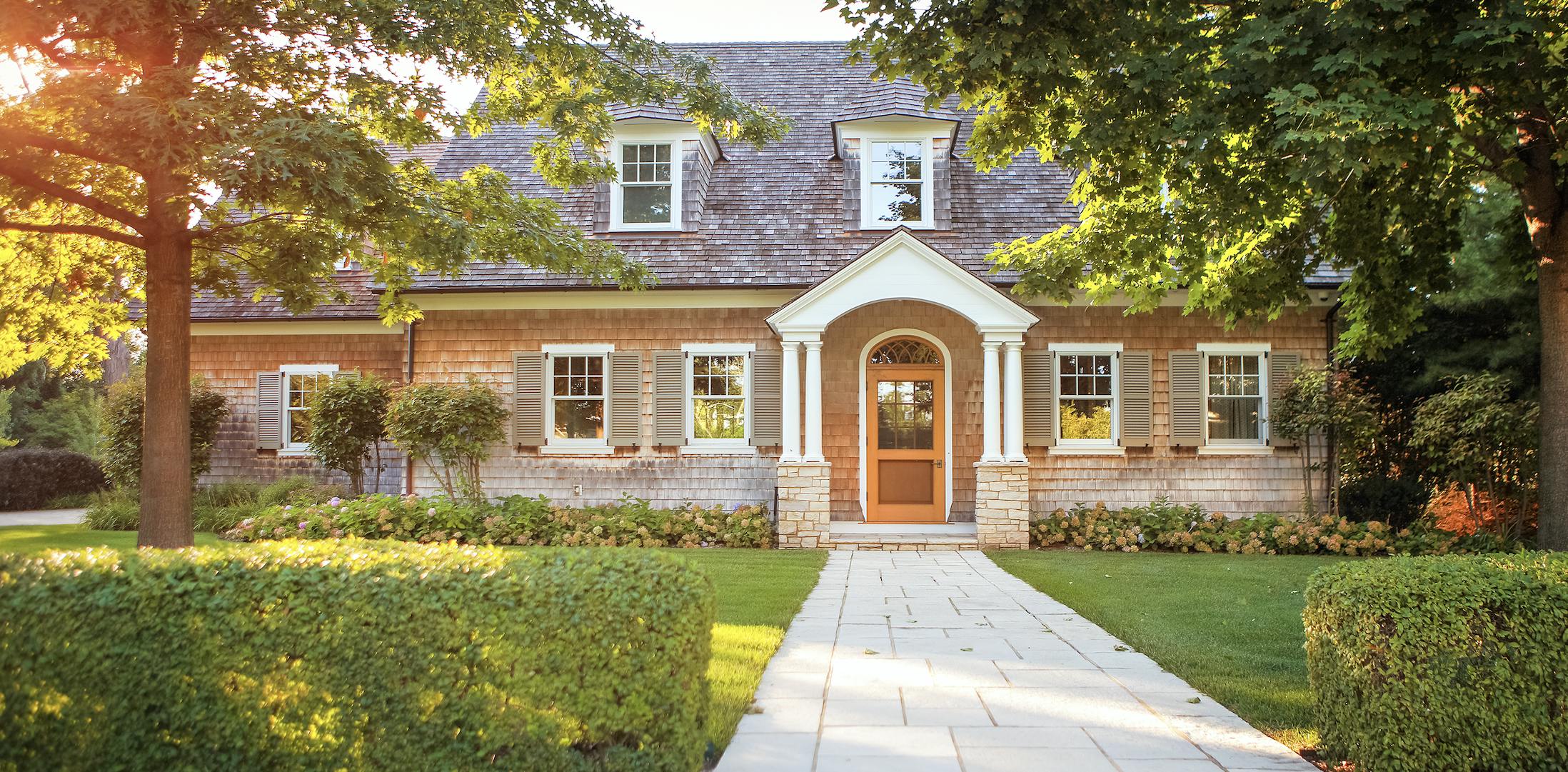 Home with shingle siding, shutters and green grass. Light is pouring in through the tree branches in the front yard.