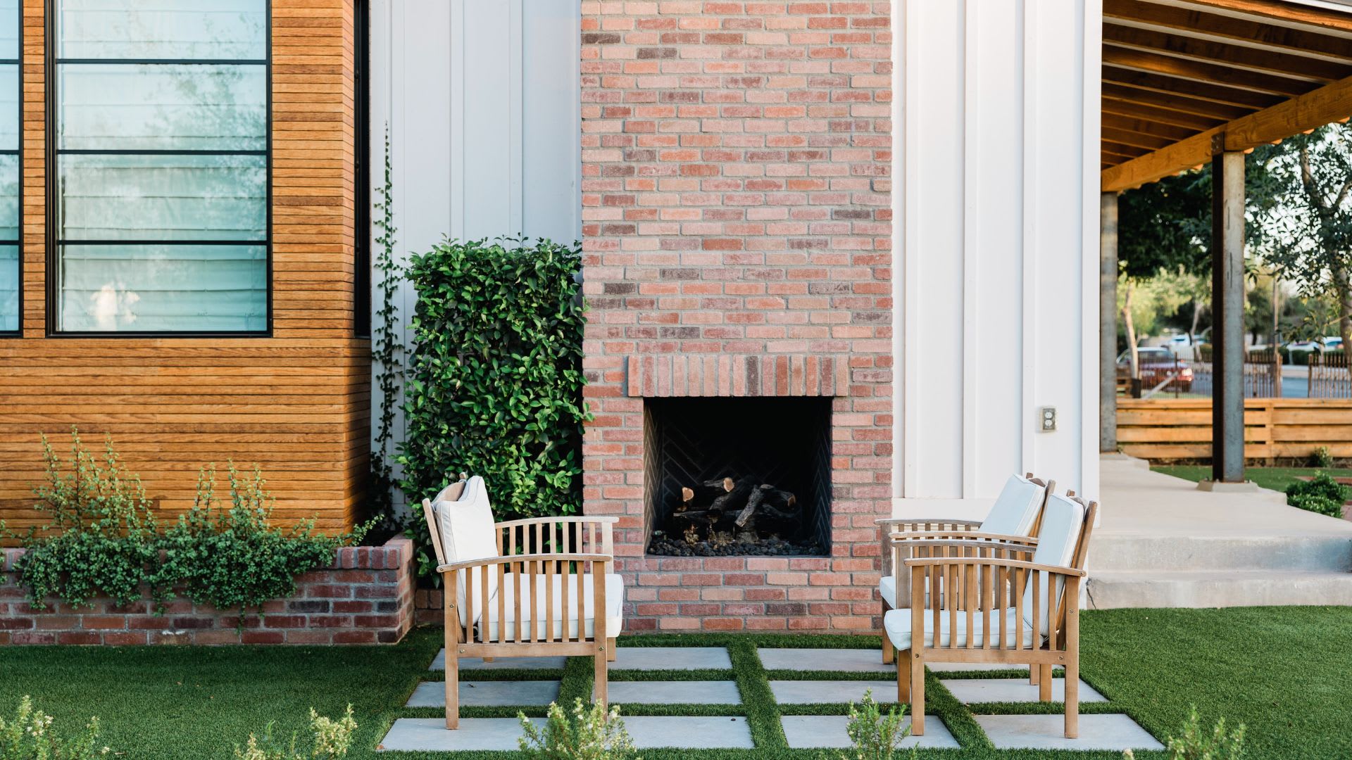 Micro escape at home with two teak chairs facing a brick outdoor fireplace surrounded by greenery and stone pavers