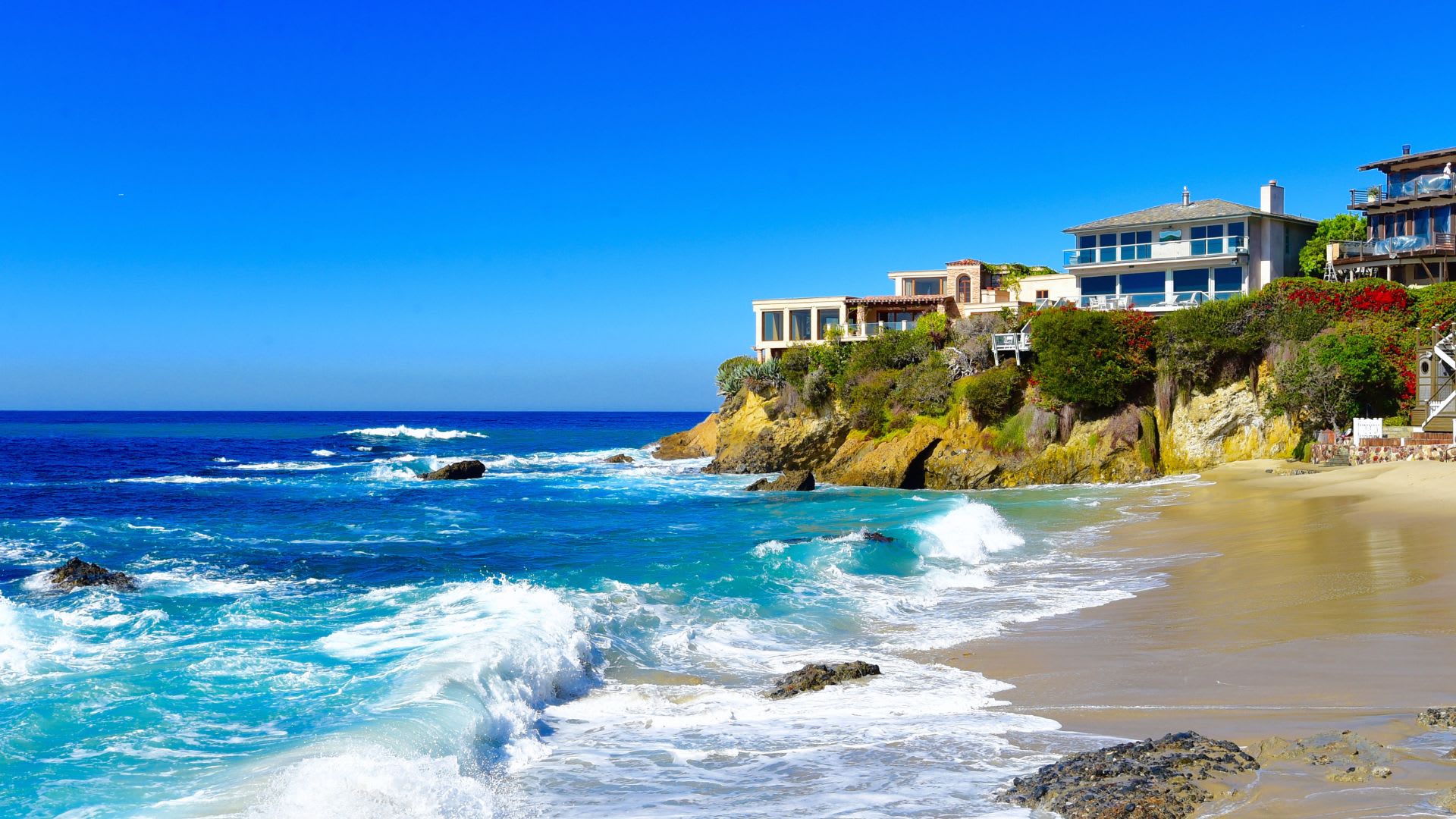 Luxury homes perched on a rocky cliffside overlooking a sandy beach with turquoise waves under a clear blue sky in Costa Rica.