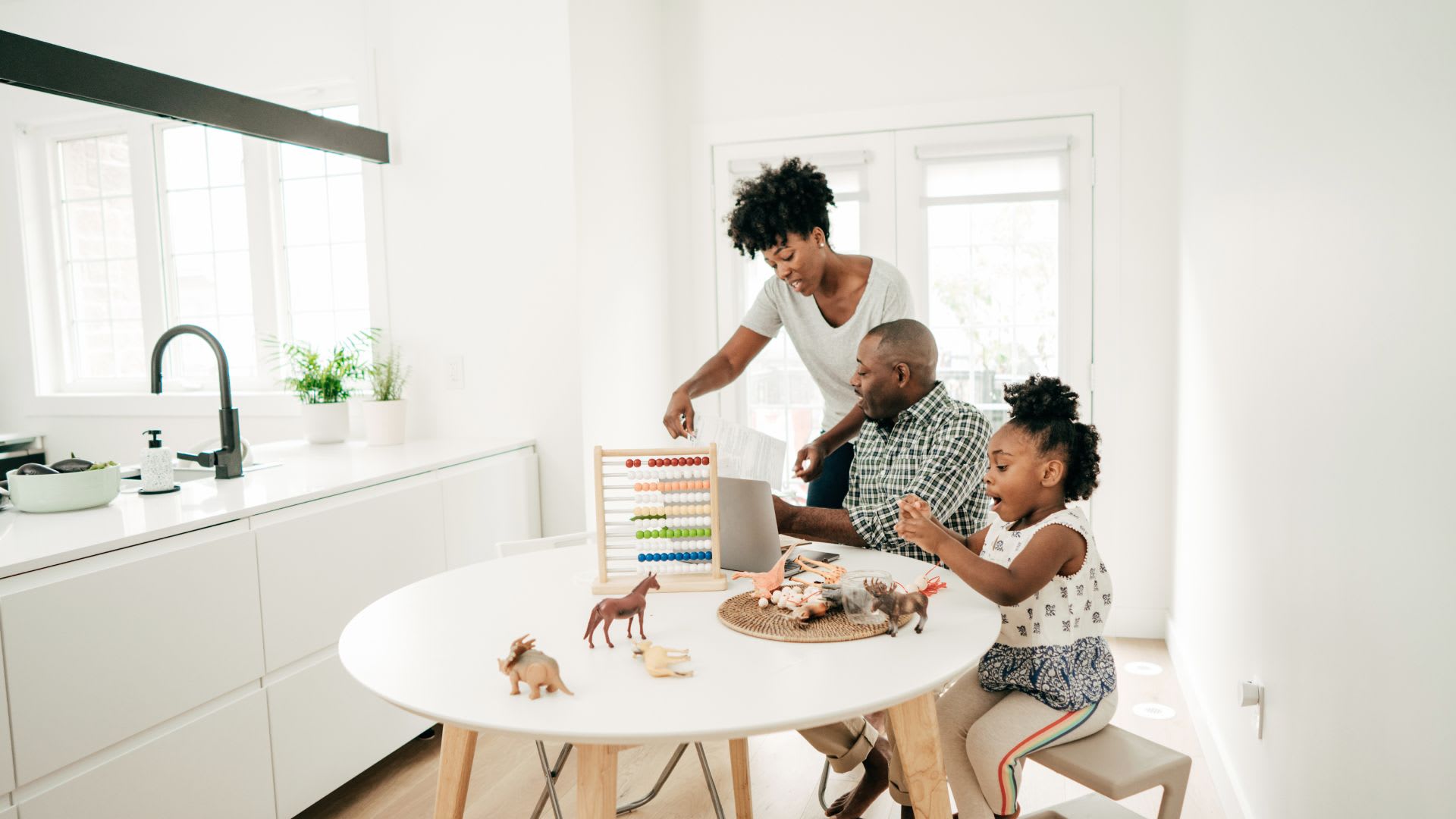 A family of three — two parents and a young child — playing with an abacus and toy animals at a kitchen table in a bright, modern white home