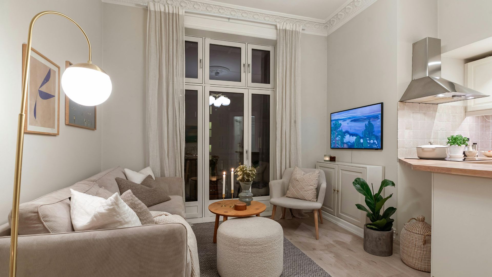 A bright, neutrally decorated living room and open kitchen in a modern home, featuring a beige sofa with throw pillows, a round wooden coffee table, a white pouf ottoman, an armchair, a wall-mounted TV, tall French doors with floor-length curtains, ornate crown moulding, and a partial view of a kitchen with a range hood and subway tile backsplash.