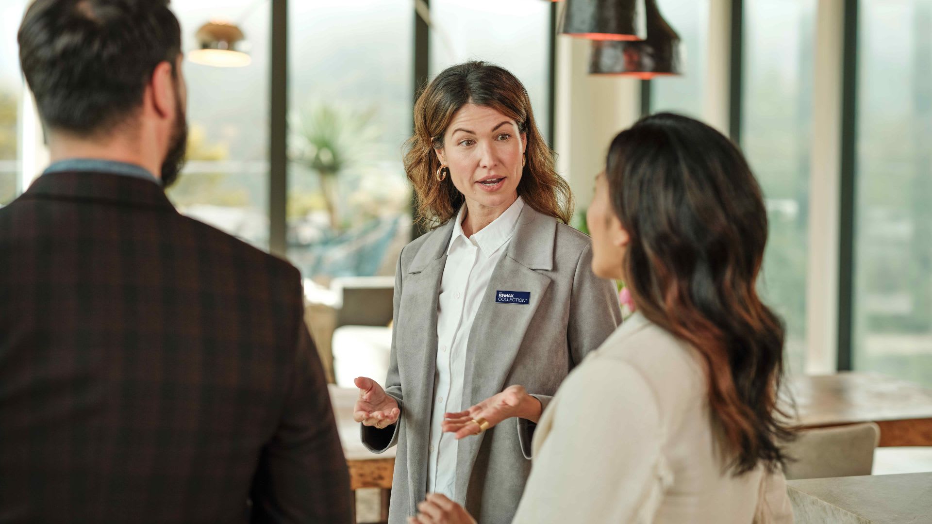 A REMAX agent in a grey blazer speaks with a couple across a kitchen island in a bright, window-lined home, illustrating why use a real estate agen