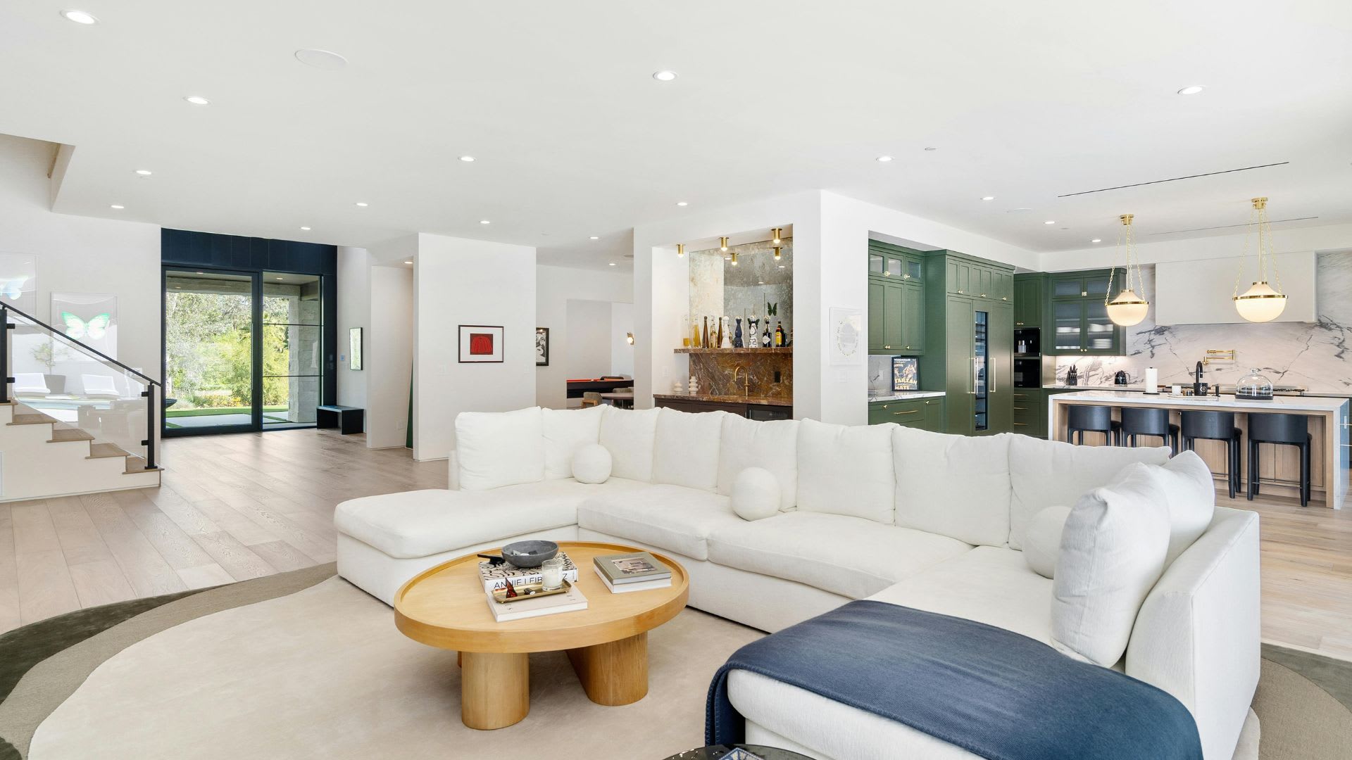 Bright, open-concept living space with a large white sectional sofa and round wood coffee table in the foreground, a kitchen with dark green cabinetry, marble countertops, and gold pendant lighting in the background, and light hardwood floors throughout