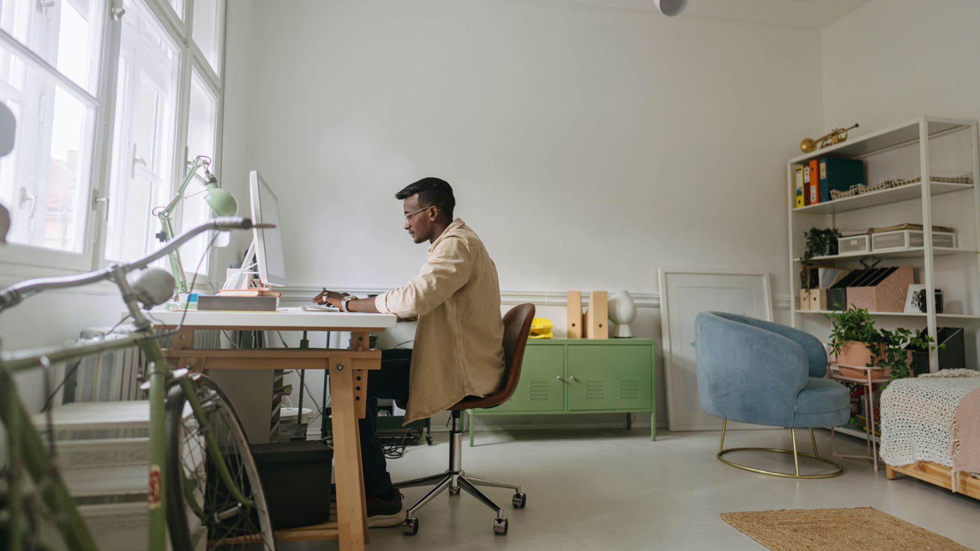 Man working at a standing desk in a bright home study space with tall windows, open shelving, and a blue accent chair