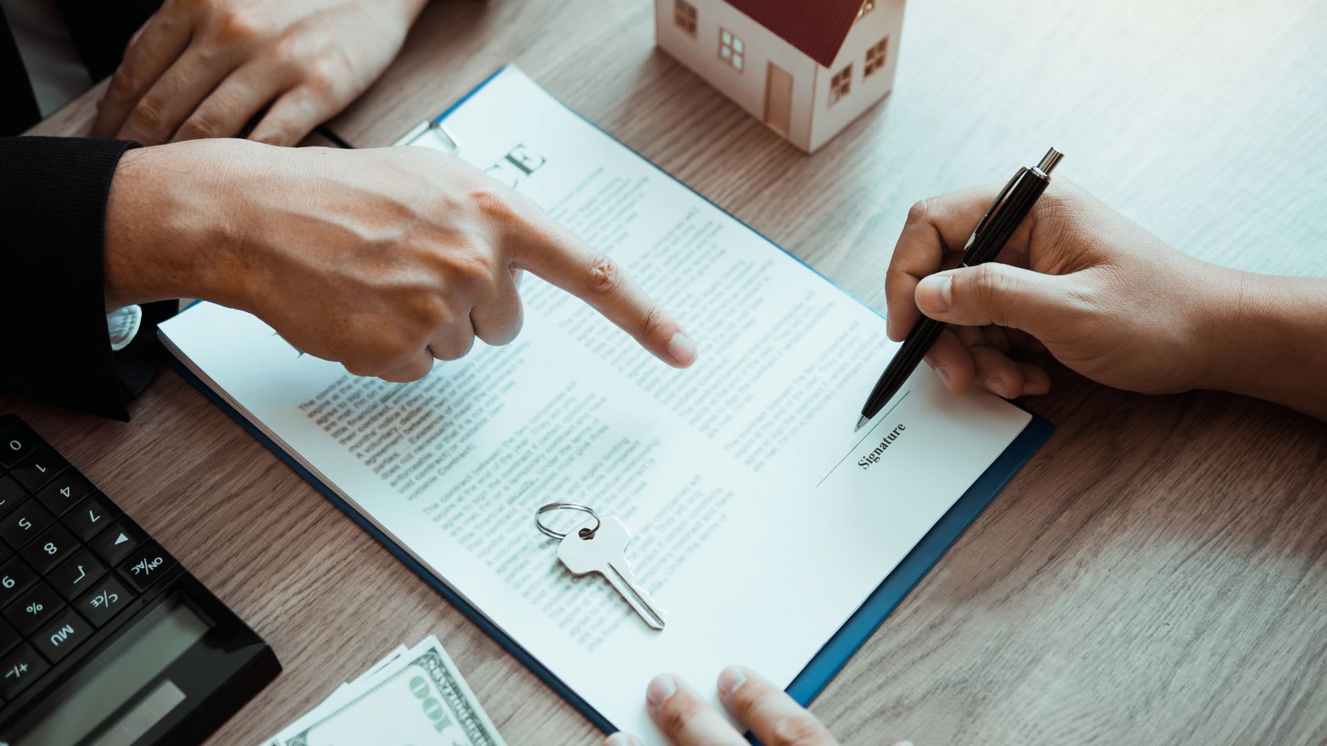 The True Cost of Buying vs. Renting A smiling professional in a tan blazer sits at a desk, gesturing while speaking with a client across the table, papers and a binder visible in the foreground.
