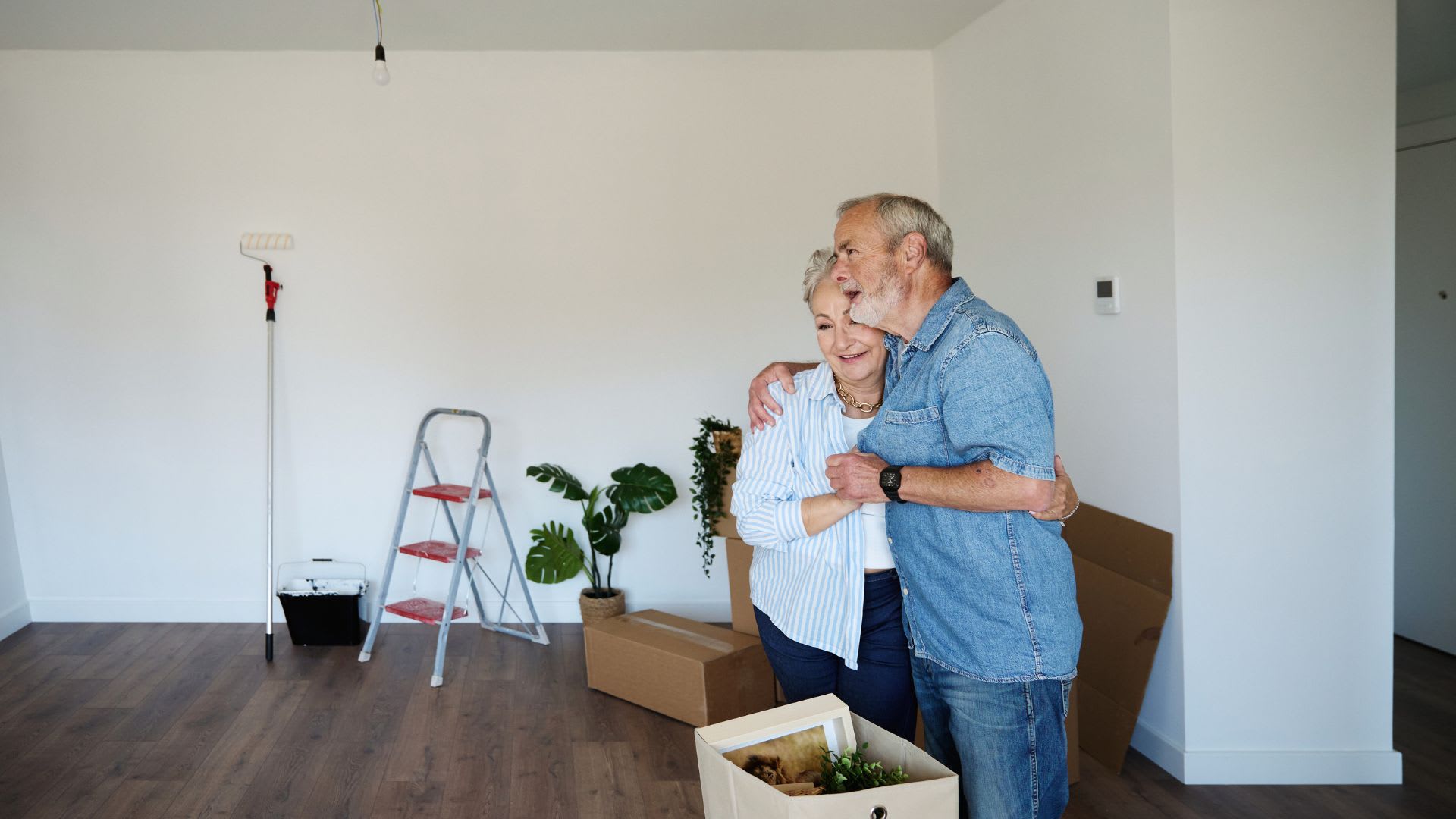 An older couple embraces while standing in an empty, unfurnished room, surrounded by moving boxes, plants, and a step ladder, taking in their new home