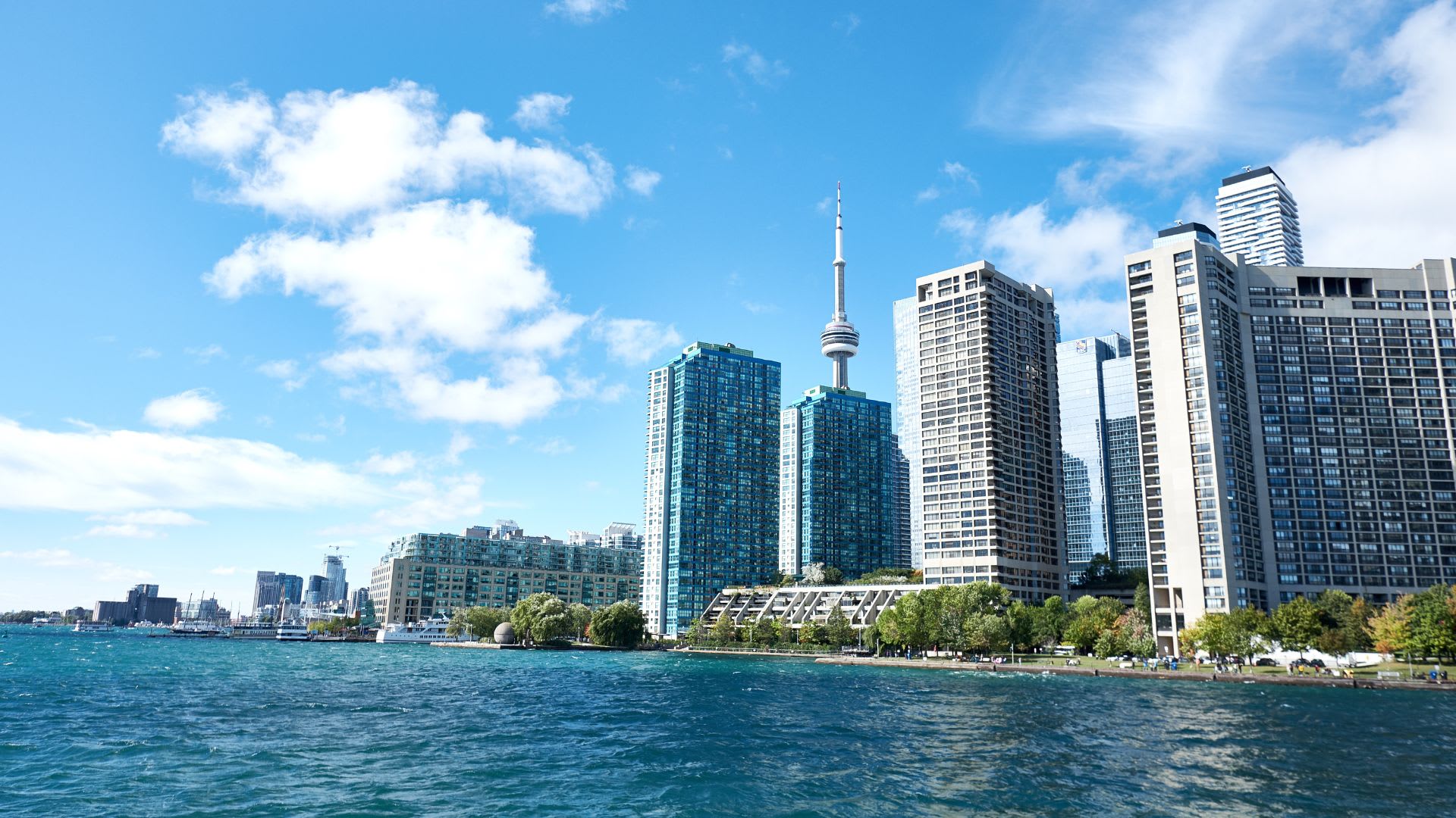 A view from Lake Ontario looking toward Toronto's harbourfront skyline, with the CN Tower rising above a cluster of high-rise condo towers along the waterfront. Green parkland with trees lines the shore in the foreground, and the water is a vivid turquoise-blue under a mostly clear sky with scattered white clouds.