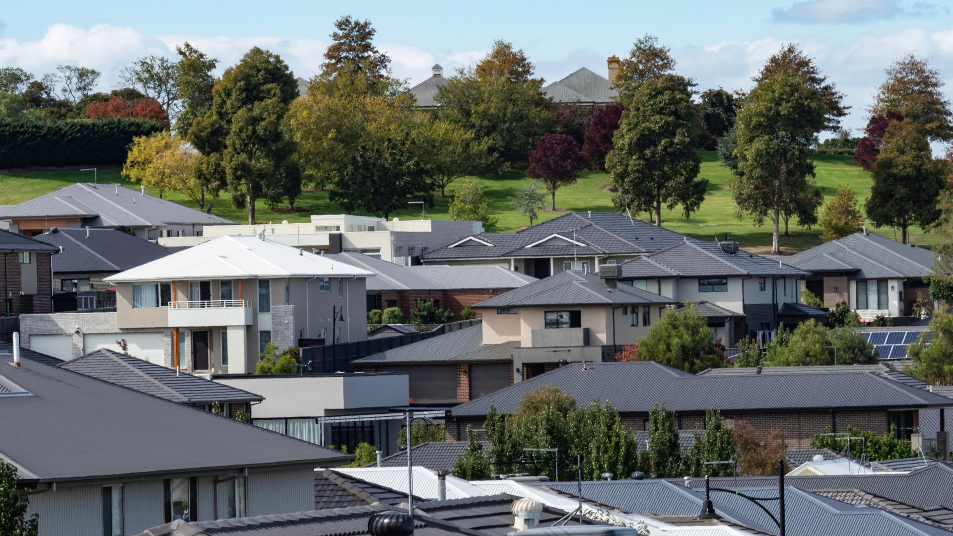 An elevated view over a modern suburban neighbourhood, showing rows of two-storey houses with grey and dark rooftops, some with solar panels, set closely together with small gardens and fences. Behind the homes, a grassy hill dotted with autumn-hued trees rises toward a large estate-style house, under a pale blue sky with light clouds.