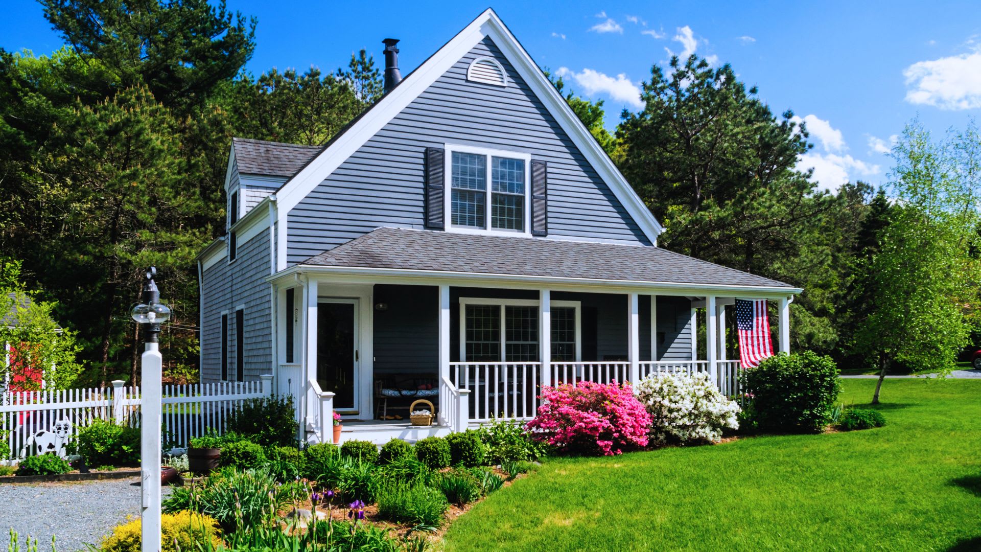 Gray Cape Cod style home with a white wraparound porch, pink and white azaleas and a green front lawn