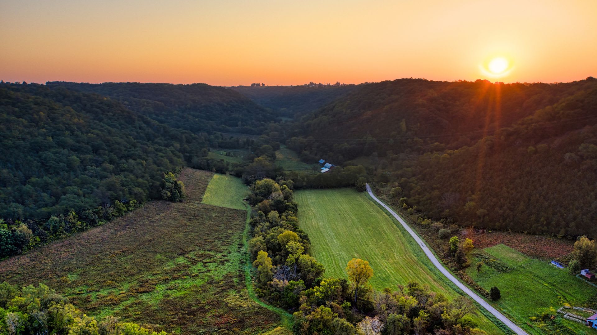 Aerial view of open fields and a gravel road running through a wooded valley at sunset