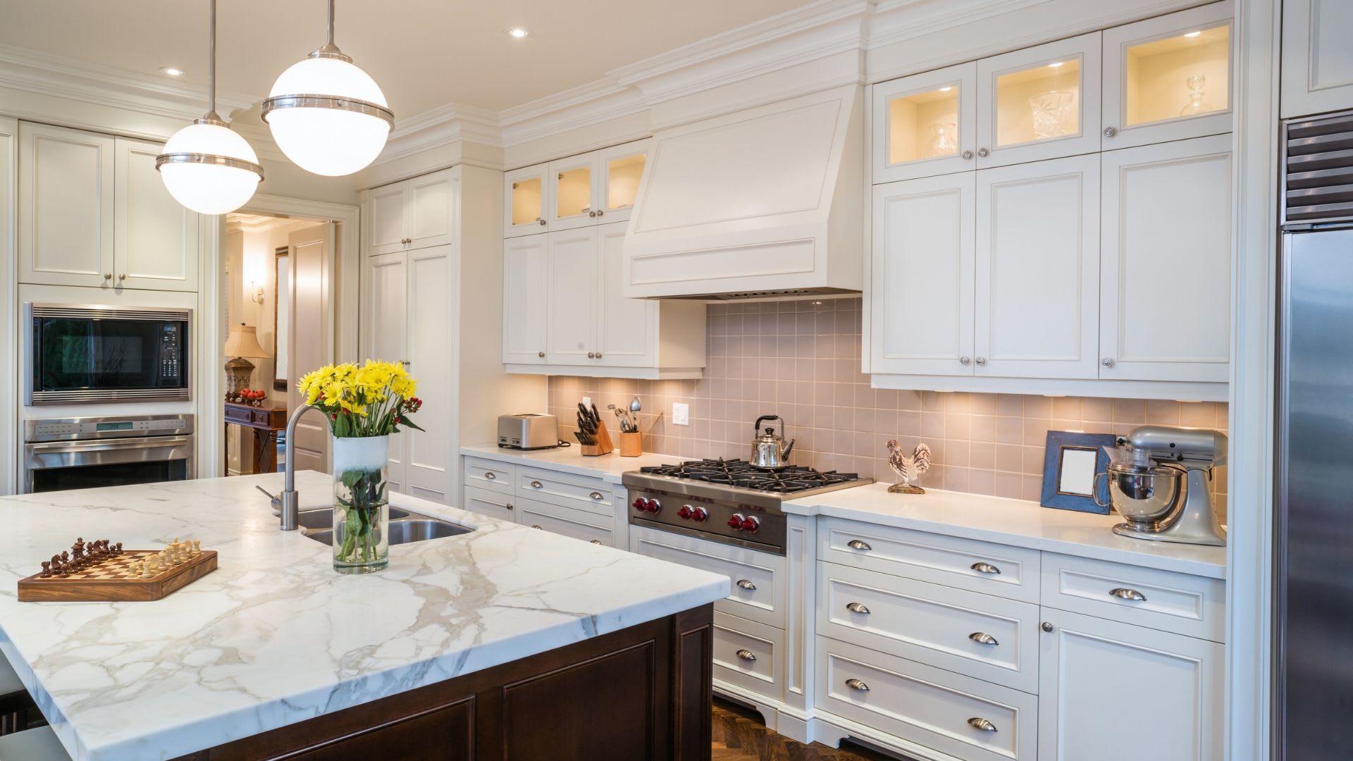 White kitchen with a marble-topped island, two globe pendant lights, a gas range with a white range hood and stainless steel appliances