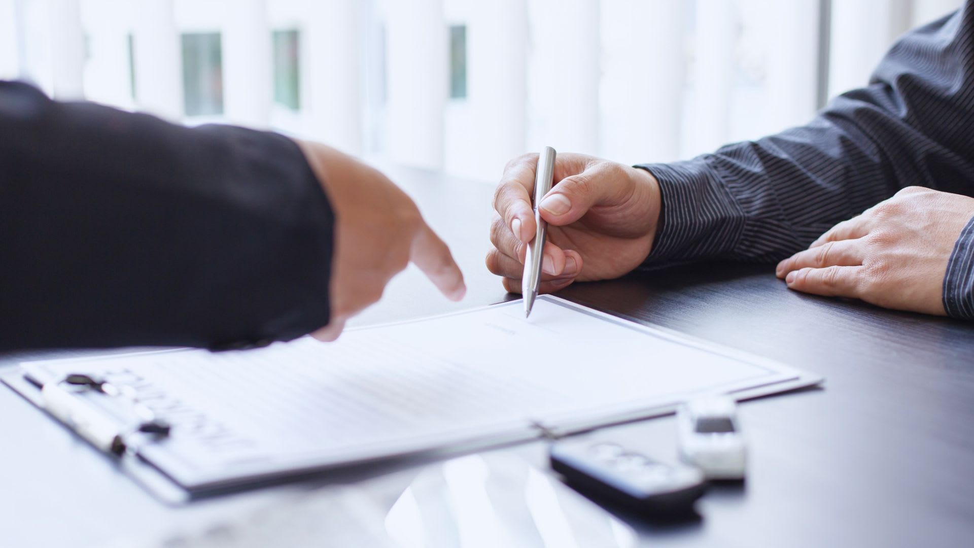 Person pointing to the signature line on a document while another person holds a pen to sign, with keys on the desk