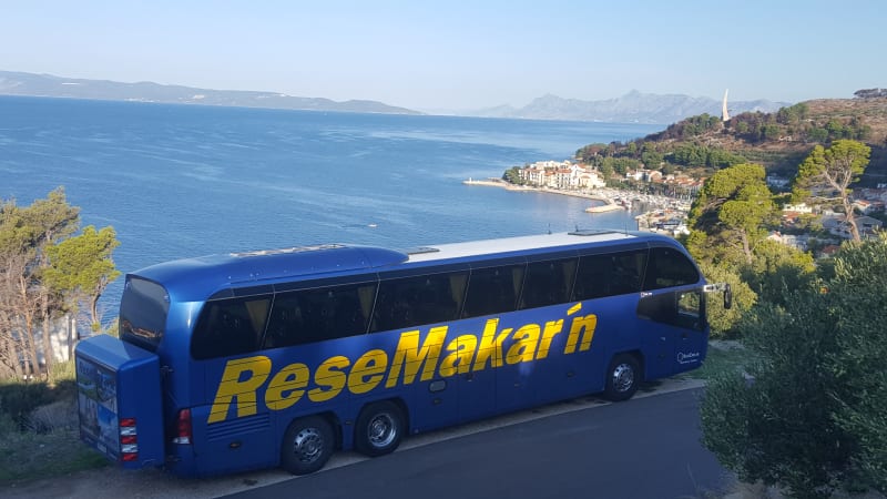 En bl buss str p en bergsvg. P bussen str det med stora gula bokstver ReseMakar n. Bakom bussen ser man ett hav.