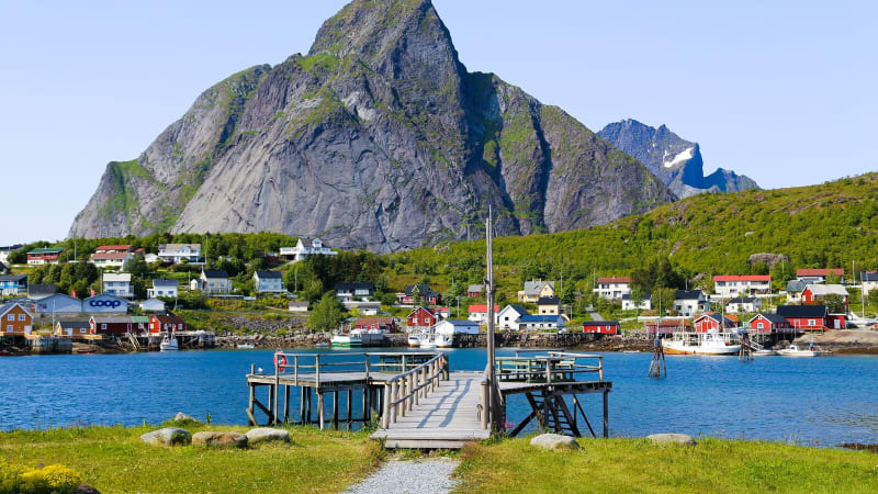 En brygga gr ut i vattnet mot en liten frgglad by. I bakgrunden reser sig ett stort berg