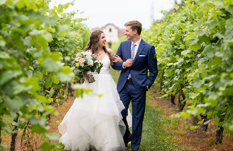 A young newlywed couple share a walk through the empty rows of a vineyard.