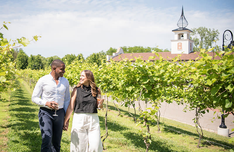 A young newlywed couple walks through a vineyard while sipping wine.